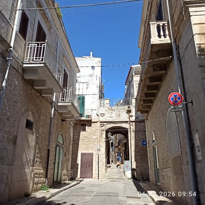 The image depicts a narrow street in Ruvo di Puglia, Italy, with stone buildings lining both sides. The street is paved and slopes gently upwards towards an arched passageway. On the left, a building features a stone facade, a wooden door, and a balcony with a white railing. On the right, another building also has a stone facade, a balcony with a decorative railing, and a circular "no parking" sign with Italian text below. A person in a black t-shirt and blue shorts is ascending a set of stone stairs through the arched passageway. The sky is clear and blue. The timestamp "06.07.2026 09:54" is overlaid on the bottom right of the image.