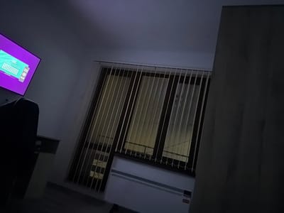The scene is an interior room located in Donchevo, Bulgaria, depicted under low-light conditions, suggesting evening or night. On the left, a flat-screen television is illuminated, displaying a purple interface featuring a keyboard layout in green and white. In the lower-left foreground, a dark, upholstered furniture piece, possibly a sofa or armchair, is partially visible. Centrally, a large window is equipped with light-colored vertical blinds, partially open, allowing external light to be discernible through and around them. Below the window, a white radiator is affixed to the wall. The right side of the frame is dominated by a tall, dark, featureless vertical surface, likely a wall or the side of a large furniture unit. No individuals are present in the observable area.