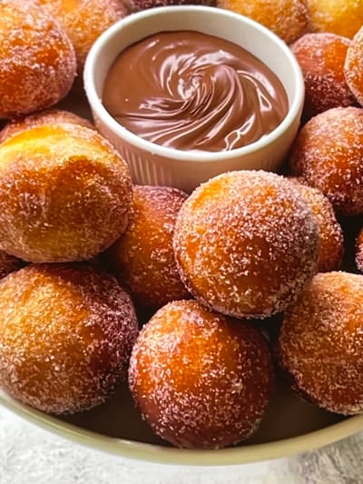 The image depicts a close-up, top-down view of a bowl filled with small, round doughnuts coated in sugar. In the center of the bowl is a smaller white ramekin filled with smooth, swirling chocolate sauce. The doughnuts are golden brown and appear to have a slightly crispy exterior, with visible sugar granules clinging to their surfaces. The lighting suggests an indoor setting, possibly a kitchen or dining area. There are no people or specific environmental details visible that would indicate a location in Nijmegen, Netherlands.