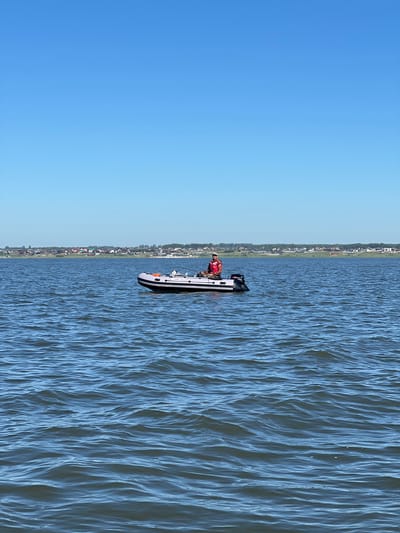 An inflatable boat with a person aboard is on a body of water. The person is wearing a red life vest and is seated in the boat, facing towards the left of the frame. The boat has a small outboard motor on the back. In the background, a shoreline with houses and trees is visible under a clear blue sky. The water is choppy with small waves. This scene is located in Novopichugovo, Russia.