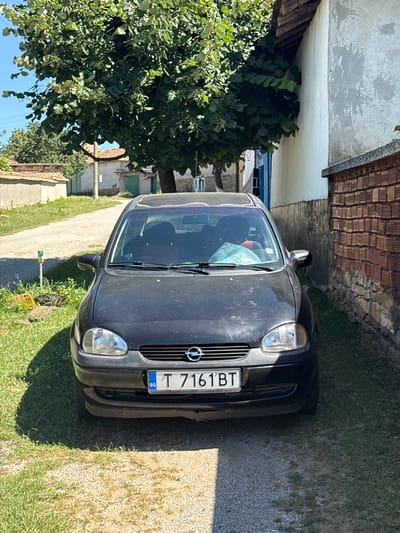 A black Opel Corsa is parked on a gravel driveway in front of a building with a brick wall. The car is facing forward, and its license plate reads "T 7161 BT" with "BG" indicating Bulgaria. A large tree with green leaves partially overhangs the car and the building. The surrounding environment appears to be a rural or village setting, with a dirt road leading away from the car and other buildings visible in the background. The lighting suggests it is a sunny day.