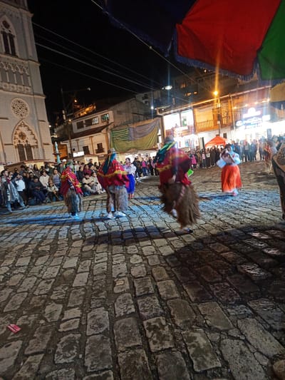 The image captures a cultural performance in El Cisne, Ecuador, at night. The scene unfolds in a public square paved with cobblestones, with a large crowd of onlookers.
Several performers are present, clad in traditional attire consisting of colorful shawls, wooly leg coverings, and headdresses. One woman wears a vibrant orange skirt and white blouse. The performers are engaged in some form of dance or ritual movement.
In the background, the illuminated facade of a church is visible. Buildings, street lights, and overhead decorations are also present. The overall atmosphere suggests a public festival or celebration.