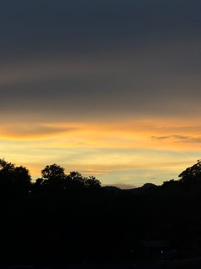 The image captures a silhouetted landscape at dusk, likely in Luang Prabang, Laos. The sky is dominated by a gradient of colors, transitioning from a deep, dark blue at the top to vibrant bands of orange and yellow near the horizon. Wispy clouds are visible within these colored bands. Below the sky, a dark, silhouetted treeline spans the width of the image, with the irregular shapes of treetops clearly defined against the lighter sky. In the distance, the silhouettes of rolling hills are visible behind the trees. The lower portion of the image is almost entirely black, obscuring any details on the ground, though faint indications of structures or lights can be barely discerned in the far lower right corner. No people or specific identifiable objects are clearly visible due to the darkness and silhouettes. The scene evokes a tranquil and natural setting during twilight.