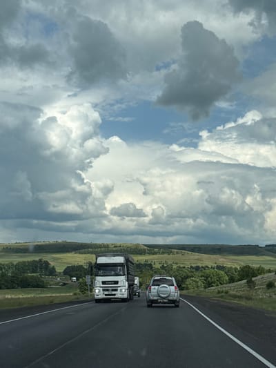 A white KAMAZ truck is driving on a paved road in Kendektamak, Russia. The truck has its headlights on. A silver SUV is driving behind the truck. The road is lined with green grass and trees. In the distance, there are rolling hills. The sky is cloudy and blue. The image was taken from inside a moving vehicle.