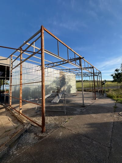 The image shows a metal framework structure, likely a greenhouse or similar agricultural building, standing outdoors. The metal is rusty in places. Behind the structure is a corrugated metal sheet partially covering the back. The ground is concrete, cracked and weathered. In the background, there is a grassy field and some trees. The sky is blue with some clouds. The location is Dabnik, Bulgaria.