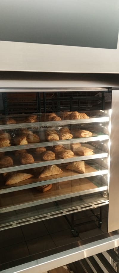 The image shows a commercial bakery display case filled with numerous golden-brown pastries. The display case has several glass shelves, each holding a variety of baked goods, appearing to be croissants, turnovers, or similar puff pastry items. The pastries are arranged neatly on brown paper liners on the shelves. The background appears to be a commercial kitchen or storefront setting, with metal shelving visible behind the display. The lighting suggests an indoor environment, possibly a bakery in Zhirnovsk, Russia, given the context provided. There are no people visible in the image. The overall impression is one of a well-stocked bakery ready for business.