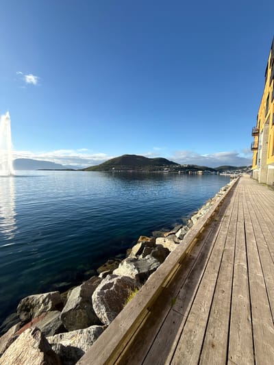 The image shows a waterside boardwalk next to a modern building in Harstad, Norway. The boardwalk is made of weathered wooden planks and runs along the right side of the frame, leading towards a body of water. The water is calm and deep blue, with ripples visible. A line of large rocks borders the water's edge. In the distance, a hill covered in green vegetation rises from the water, with buildings visible on its slopes and summit. A large fountain is visible in the water on the left side of the image, spraying a tall plume of water. The sky is a clear, bright blue with a few scattered white clouds. The building on the right is a multi-story structure with yellow siding and balconies, suggesting it is residential or commercial.