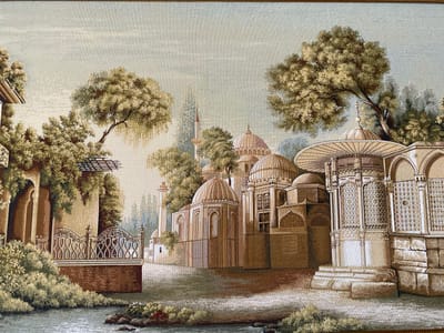This tapestry depicts a serene architectural scene with a body of water in the foreground. The focal point is a complex of ornate buildings with domes and minarets, suggesting a mosque or a similar religious or historical structure. Lush trees with dense foliage surround the buildings, with one large tree dominating the upper right portion of the image. A decorative fence with intricate patterns is visible on the left, bordering the water and a paved area. The architecture features arched doorways and windows, with detailed stonework and metallic accents on some elements. A slender minaret with a pointed spire rises from behind the main structures. The sky is a pale, hazy blue with subtle cloud formations, indicating a tranquil day. The lighting suggests it could be late afternoon or early morning, with a soft, diffused light casting gentle shadows. The overall impression is one of historical grandeur and peaceful contemplation. The scene is rendered in a rich palette of browns, beiges, greens, and muted blues, giving it a timeless quality. No people are visible in the image, and no text is present. The style suggests a depiction of a Middle Eastern or Mediterranean setting, potentially inspired by historical architecture. Given the context of Babayurt, Russia, this tapestry might represent a historical or culturally significant site within the region or a more generalized representation of Islamic architecture.