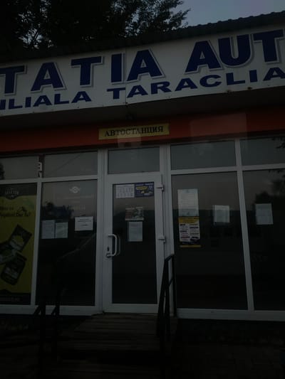 The image shows the exterior of a building in Taraclia, Moldova. The building has a large sign with blue lettering that reads "TATIA AUT JILIALA TARACLIA". Below this, a yellow sign with Cyrillic text, possibly indicating a bus station, is visible. The entrance to the building features glass doors and windows. Various posters and notices are affixed to the glass doors and the interior walls visible through the windows. A wooden ramp leads up to the entrance. The lighting suggests it is dusk or early morning, with shadows cast on the ground. No people are visible in the image.