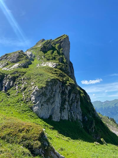 The image shows a steep, grassy mountain slope with a prominent rocky peak. A single hiker, wearing dark clothing and a backpack, is visible ascending the slope on a narrow path on the left side of the frame. The sky is clear blue with some wispy white clouds. In the distance, other mountain ranges can be seen. The sun is shining brightly, casting shadows on the mountain. The location is described as Brienz (BE), Switzerland, suggesting this is a mountainous area near the town.