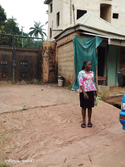 A woman in a patterned blouse and black skirt stands in a dirt area near a building. The building is under construction with open window frames and a covered shed. Behind the woman is a gate and some trees are visible in the background. A blue car is partially visible in the lower right corner. The ground is uneven and has tire tracks. The image is somewhat underexposed.