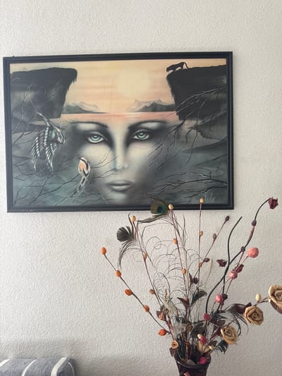 The image displays a framed artwork hanging on a textured white wall. The artwork is a surrealist painting featuring a large human face emerging from a landscape. The face has striking green eyes and is surrounded by dark, rocky cliffs, a body of water, and a misty horizon. Two birds and a silhouette of a panther on a cliff are also depicted in the painting. In the foreground, a vase filled with dried floral arrangements, including feathers and other decorative elements, sits on a surface below the painting. The location is identified as Belo pole, Bulgaria, although no specific landmarks or contextual clues within the image confirm this.