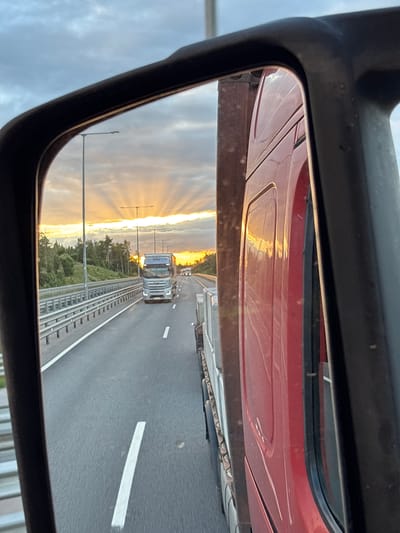 The image shows a view from a truck's side mirror, reflecting a highway at sunset. In the reflection, a silver truck is driving on the highway, with trees and streetlights visible on the side. The sky is dramatic with sun rays breaking through clouds. The foreground of the image shows the side of a red truck. The scene is set on a highway, likely in or near Lapustino, Russia, given the context.