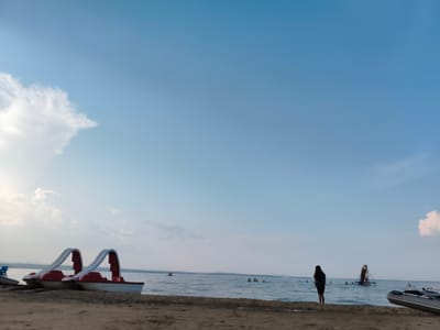 The image depicts a beach scene on a sunny day. In the foreground, two red and white pedal boats are parked on the sand. A single person stands on the shore, facing the water, with several other individuals swimming or wading in the distance. Further out in the water, there is a floating structure with a slide. The sky is a clear blue with a few scattered clouds. An inflatable boat is partially visible on the right side of the frame. The scene appears to be the beach area of Gubernskoe, Russia, likely during a recreational period.