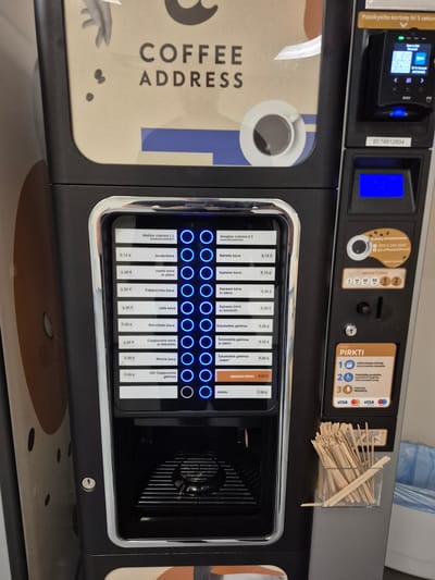 This is a close-up, eye-level, full-shot photograph of a coffee vending machine in Pabradė, Lithuania. The machine is black with a metallic silver trim around the screen and the dispenser area. The machine features a large screen displaying various coffee options and prices in Lithuanian, with selectable buttons next to each option. A card reader is visible on the upper right side, with a blue display screen below it. There is a slot for coins and a compartment for stirrers on the right side of the machine. The machine is situated in an indoor public space, likely a transit hub or a communal area, given the presence of other vending machines and the general aesthetic. No people are present in the image, and the focus is solely on the vending machine and its immediate surroundings.