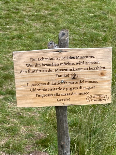 The image shows a wooden sign with a message in both German and Italian. The sign is attached to a wooden stake, which is planted in the ground. The ground surrounding the stake is covered in short green grass. The sign reads: “Der Lehrpfad ist Teil des Museums. Wer ihn besuchen möchte, wird gebeten den Eintritt an der Museumskasse zu bezahlen. Danke! Il percorso didattico fa parte del museo. Chi vuole visitarlo è pregato di pagare l'ingresso alla cassa del museo. Grazie!” Below the Italian text, the sign is stamped with the word "PLATNER" in a stylized font and the word "Bienenhof". The sign is located in the town of Costalovara, Italy, at coordinates 46.525° N, 11.428° E.