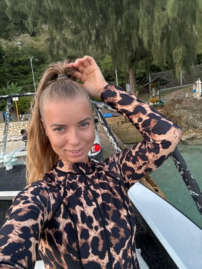 A blonde woman with blue eyes smiles at the camera, adjusting her high ponytail. She is wearing a long-sleeved leopard print rashguard. She is on a boat, with the ocean and a rocky, vegetated shoreline in the background. A car is visible parked near the shore. The lighting suggests it is either early morning or late afternoon. The location is Christmas Island, an Australian territory in the Indian Ocean, approximately 350 kilometers south of Java and 1,300 kilometers northwest of the Australian mainland.