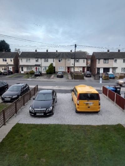 The image captures an overhead view of a residential street, looking down into a driveway and beyond to a row of terraced houses under an overcast sky. The scene is oriented as if rotated 90 degrees counter-clockwise, showing the sky on the left, a line of houses and a street in the middle, and a gravel driveway with a lawn on the right.
In the foreground (right side of the rotated image), a light grey gravel driveway extends from the bottom of the frame towards the top. A patch of green grass borders the driveway on its right side. Two black cars and one yellow van are parked on the gravel. Closest to the viewer is a black Audi, partially visible, with the license plate "AU60 JUU". Further along the driveway is a black Mercedes-Benz sedan, license plate "P16 LJJ". A bright yellow van, possibly a Ford Transit Connect, is parked next to the Mercedes, facing towards the top of the frame, displaying the license plate "GTIP 08J".
Beyond the driveway, a paved road or street runs horizontally across the middle of the frame. Several more vehicles, including a blue car and other dark-colored vehicles, are parked along this street. On the far side of the street (left side of the rotated image), a row of uniform terraced houses extends into the distance. These houses have varying front doors (red, white, brown) and appear to be two or three stories high, with pitched roofs. Some have small front gardens or paved areas. Power lines are visible strung between poles and across the sky, particularly on the far left.
The sky is cloudy and grey, suggesting an overcast day. No people are visible, and there is no apparent activity taking place other than the static presence of vehicles and buildings. The overall impression is that of a quiet, typical suburban street in the daytime.