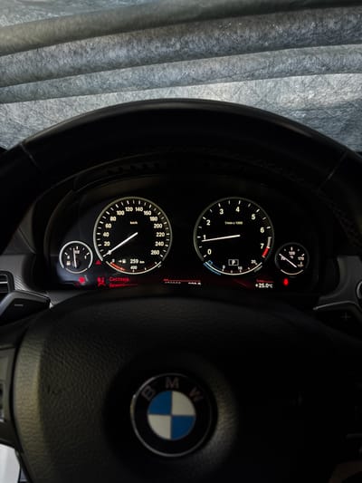 This media file depicts the interior of a car, specifically focusing on the dashboard and steering wheel. The subject is the car's instrument cluster, which displays various readings like speed, engine RPM, fuel level, and outside temperature. The car's brand, BMW, is visible on the steering wheel emblem. The scene is dark, suggesting either nighttime or a dimly lit interior. The dashboard lights are illuminated, providing a futuristic glow to the gauges. There are no people visible in the image. The outside temperature reading of +25.0°C indicates a warm day. The text visible includes "km/h," "1/min x 1000," "Temp.," "Система," and "безопаснос," which is Russian for "System" and "safety." The location context provided is Novy, Russia. The overall impression is one of modern automotive technology within a private vehicle.