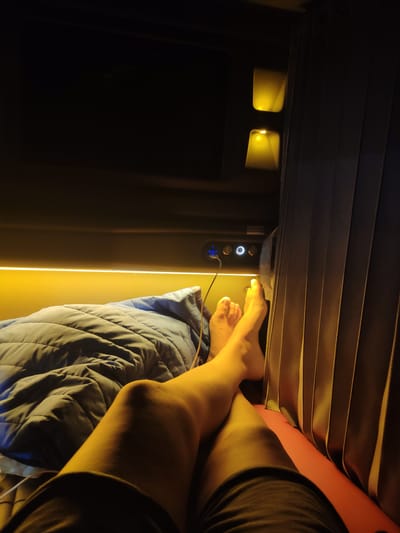 The image depicts a person's legs and feet resting on a padded surface, possibly a bed or a seat, within a dimly lit, modern capsule hotel or similar accommodation. The person is covered by a blue duvet. The environment is characterized by warm, yellow ambient lighting, with built-in light fixtures visible in the upper portion of the frame. A control panel with illuminated buttons and a USB port is located to the left of the person's feet. The wall to the right features vertical pleats, suggesting a curtain or textured panel. The scene appears to be in a private sleeping pod. The location is stated to be in M'Drắk, Vietnam.