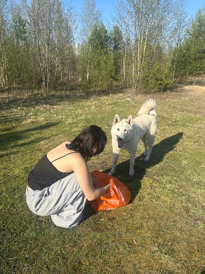 The image shows a woman sitting cross-legged on a grassy field. She is wearing a black tank top and gray sweatpants, and her back is facing the camera. An orange plastic bag is near her. A white dog is standing near her, with its tongue out. The field is partially shaded and there are trees in the background. The location is in the city of Vysokii, Russia.