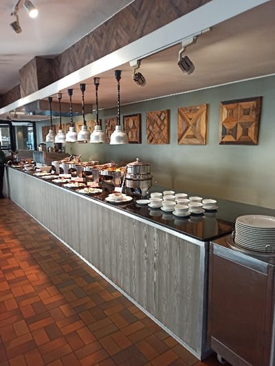 A sideways interior shot reveals a buffet arrangement with stacked plates and chafing dishes atop a gray wooden counter. Above, a wall adorned with four decorative wood panels leads to a series of gooseneck lamps. To the right, a reddish-brown brick floor is visible. The scene is illuminated, highlighting the textures of wood and brick.