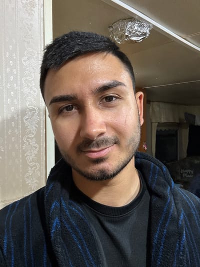 The image displays an adult male subject centered within an indoor setting, identified as Muirhead, United Kingdom. The individual has a light-to-medium skin tone, short dark hair, dark eyebrows, and brown eyes, presenting a faint scar on his right cheek and light facial hair on his chin and jawline. He is captured looking directly at the camera with a subtle smile. Clothing consists of a black long-sleeved shirt overlaid by a dark blue fleece robe featuring black vertical stripes. A thin gold chain is visible at his neck. The immediate background on the left is a wall covered with light-toned, patterned wallpaper displaying a repeating floral or scrollwork design, adjacent to a vertical light-colored trim. The ceiling, light in color, has a crumpled piece of aluminum foil affixed to it above the subject's upper right head. The background on the right is darker and less distinct, revealing an interior space with a draped light-colored fabric and a partially visible sign that reads "Happy Place."