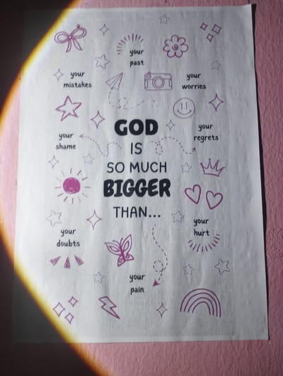 This media file is a close-up, eye-level shot of a piece of paper with a handwritten message and drawings, possibly a poster or a page from a notebook. The paper is light-colored and appears to be textured. It is placed against a textured pink background, suggesting it's mounted on a wall or displayed on a surface. A diagonal band of warm, yellow-orange light falls across the left side of the paper, indicating artificial lighting or a light source positioned to the left and slightly above the frame.

The central text on the paper is prominently displayed in bold, dark letters: "GOD IS SO MUCH BIGGER THAN...". Surrounding this central message are various small, purple drawings and accompanying text. These drawings are simple and symbolic, including a bow, stars, a paper airplane, a camera, a smiley face, a flower, a sunburst, a crown, hearts, a butterfly, a rainbow, and a lightning bolt. Each drawing is paired with a word or phrase written in a smaller font, such as "your past," "your mistakes," "your worries," "your shame," "your doubts," "your regrets," "your hurt," and "your pain." These elements suggest a theme of overcoming personal struggles or negative emotions through faith.

The overall impression is one of a personal or inspirational message, likely created as a reminder or affirmation. The lighting creates a dramatic effect, highlighting the texture of the paper and the pink background. There are no people visible in the image. The activity taking place is the display of this handwritten message. The location context of Iyafin, Nigeria, places this within a specific geographical and cultural setting, though the visual elements themselves do not provide direct cues to this location. The emotions conveyed are those of hope, reassurance, and perhaps a sense of spiritual strength.