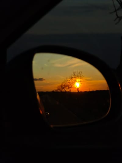 A sunset is reflected in a car's side mirror. The sky is a mix of orange and yellow hues. A line of bare trees stands silhouetted against the bright sun. A dark road stretches toward the horizon. The surrounding area outside the mirror is dark, suggesting the interior of the car at night. The mirror's frame is visible, confirming it is a side mirror. There is no specific information that identifies the image as taken in Vaglen, Bulgaria.