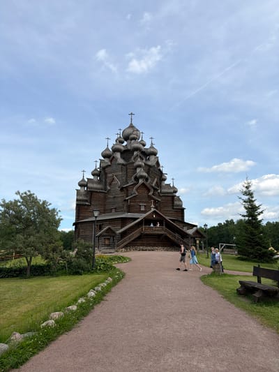 In Nevskii Parkleskhoz, Russia, a large, intricate wooden church with numerous domes and crosses stands prominently against a bright blue sky with scattered white clouds. The church, constructed of dark wood, features multiple levels and ornate architectural details. In front of the church, a gravel path leads towards the entrance, bordered by a lush green lawn and a line of rocks. Several people are walking on the path, appearing to be tourists. To the right of the path, a tall evergreen tree stands, and further on, a rustic wooden bench is visible. The surrounding environment is natural, with trees and greenery indicating a park or open space setting.