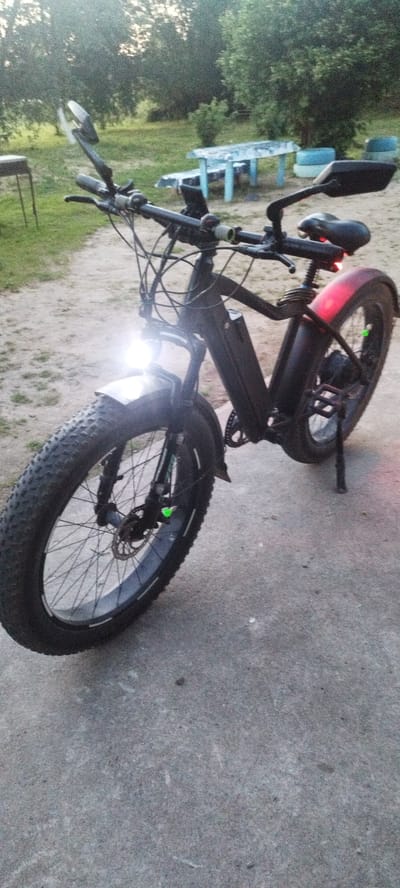 The image shows a black fat-tire electric bicycle parked outdoors on a paved surface, likely a path or road. The bicycle is the primary subject. It has thick tires, a prominent battery pack mounted on the frame, a bright headlight that is currently on, and a red taillight. The handlebars are equipped with mirrors. In the background, there is a dirt path, grass, and trees, suggesting a rural or park-like setting. A picnic table with blue benches is visible in the middle ground, and further back, some blue tires are stacked. The lighting suggests it is late afternoon or early evening, with the sun low in the sky, casting a warm, diffused light. The weather appears to be calm and clear. There are no people visible in the image, and no discernible text. The scene conveys a sense of readiness for a ride or a moment of rest during an outdoor excursion. The location context of Сінькоўскі сельскі Савет, Belarus, indicates a rural area in Belarus.