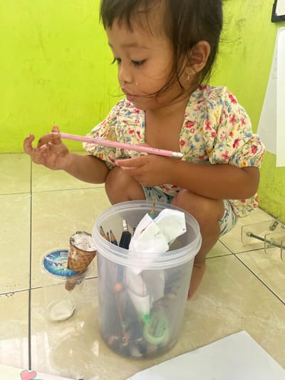 An indoor scene in Desa Batu Layar, Indonesia, depicts a young child squatting on a light-colored tiled floor with dark grout lines, in front of a bright lime green wall. The child has dark hair, wears a floral-patterned short-sleeved top with a cream background and pink, blue, and yellow flowers, and patterned shorts. A small gold earring is visible in her right ear. White residue is present on the left side of her upper lip and cheek. Her hands, particularly the left, are smudged with a dark substance. She holds a pink pencil with a white eraser tip in both hands, looking downwards with a focused expression.
To her left, on the floor, is a clear plastic cylindrical container filled with various pencils (some sharpened, some unsharpened, some with erasers), pencil shavings, a green pencil sharpener, and crumpled white paper. Immediately to the left of this container, a plastic cup holds an ice cream cone topped with chocolate chip ice cream. The cup has a blue and white label partially visible. An empty, clear plastic cup is positioned next to it. In the background on the right, partially visible white sheets of paper are attached to an unseen surface, alongside a clear acrylic stand or structure.