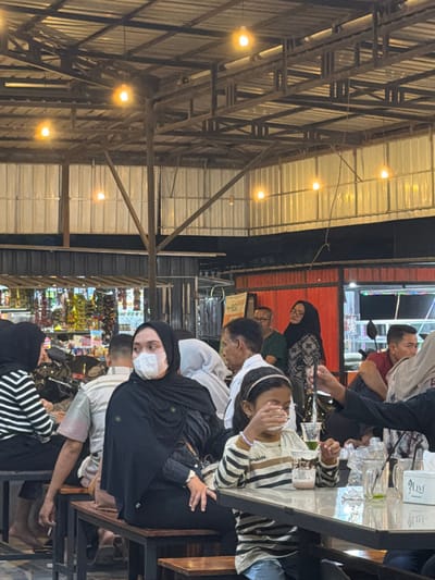 The image depicts an evening scene within an indoor or semi-outdoor dining establishment, likely a cafe or food court, located in Beurandang, Indonesia. The venue features an exposed structural ceiling composed of metal trusses supporting a corrugated metal roof, from which multiple bare incandescent light bulbs are suspended, providing illumination.
In the foreground, a young female child, wearing a striped long-sleeve shirt, jeans, and a silver headband, is seated at a dark-colored rectangular table. She holds a clear plastic cup containing a light-colored beverage to her mouth with both hands, actively drinking. Beside her cup on the table is another clear plastic cup holding a green liquid with a black straw, a clear plastic water bottle, and a white tissue dispenser branded "LIVI." To the child's left, a woman is seated on a wooden bench, wearing a black hijab, black clothing, and a white disposable face mask, looking slightly towards the right.
The midground and background are populated by numerous individuals, adults and possibly children, seated at similar tables and benches or standing. Visible individuals include a person in a black-and-white striped shirt and black hijab on the far left, partially obscured. A man in a white collared shirt and another man wearing glasses and a dark green shirt are visible in the mid-background. A woman in a patterned hijab is also observed further back, looking towards the right.
The environment includes dark wooden benches and dark-topped tables. In the far left background, a retail display or shelving unit holds various packaged goods. Red-painted horizontal metal beams or panels are visible in the mid-background, along with a partial sign displaying "BSI." The overall scene portrays a bustling social or dining gathering.