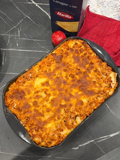 A close-up, high-angle shot shows a freshly baked pan of lasagna on a dark gray, marble-patterned countertop. The lasagna is golden brown and bubbly on top, with visible layers of pasta and meat sauce. To the left of the lasagna is a box of Barilla lasagna noodles, and to the right is a crumpled red cloth. A small, red, decorative egg with a cartoon cat face is placed near the box of noodles. The background is out of focus, but suggests a kitchen environment. There is no visible indication of the city of Lenina, Russia.