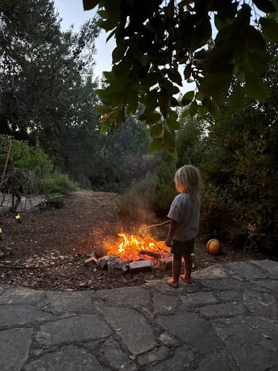 A young child with blond hair, wearing a gray striped t-shirt and dark shorts, stands on a stone patio facing a campfire. The child is holding a stick with a marshmallow over the fire. The campfire is in a pit made of rocks and surrounded by dirt and leaves. Behind the child is a dense green bush and a yellow and black soccer ball. The scene is set in an outdoor environment with trees and foliage visible in the background. The time of day appears to be dusk or early evening. The location is identified as Kfar Vradim, Israel.