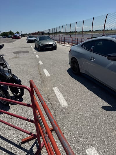 In Kaloyanovo, Bulgaria, on a bright, sunny day, a race track event is underway. A matte grey BMW M4, with license plate CB 9668 OM, is parked in the foreground on the asphalt. To its right, a silver coupe is also present. In the background, a motorcycle and a red barrier are visible, along with a chain-link fence that encloses the track. The sky is clear blue, and the sun casts long shadows across the pavement. A few tents and flags suggest a gathering or event is taking place.