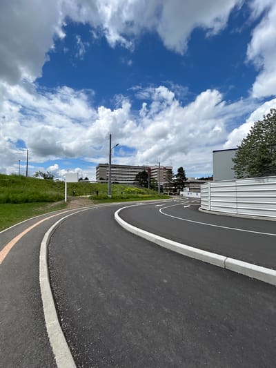 The image shows a curved asphalt path with a white border on the right and a yellow line further to the left, likely a designated bicycle path. To the right of the path is a grassy area. In the background, buildings are visible against a cloudy sky. The location is Saint-Priest-en-Jarez, France.