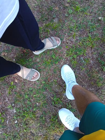 This image shows the lower bodies and feet of two individuals standing on a patch of grass and dirt. One person is wearing black pants and grey sandals, with a white shirt visible at the top left of the frame. The other person is wearing white sneakers with white socks and green shorts. The ground is covered in grass, dry leaves, and small stones, suggesting an outdoor, natural setting. The lighting appears to be natural daylight, likely daytime given the brightness. There is no discernible text in the image. The image captures a casual, everyday moment of people outdoors, possibly taking a walk or break.
