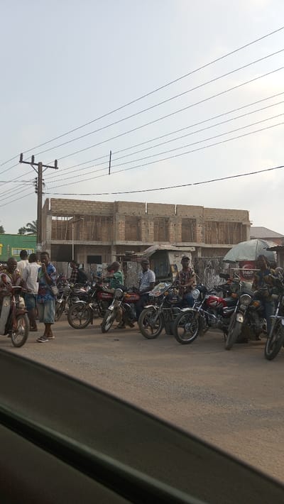 The image depicts a street scene in Ihiala, Nigeria, under an overcast or hazy sky. The perspective is from a vehicle, with a dark window frame visible in the lower left foreground. A paved road surface occupies the lower half of the frame.
In the midground, approximately fifteen adult males are present, predominantly gathered around a line of motorcycles parked or idling along the roadside. Many men are seated on their motorcycles, facing generally towards the viewer or to the right. One man in a patterned shirt and blue shorts stands holding a phone to his ear, facing left, near a red motorcycle. Another man in a white long-sleeved shirt and dark trousers stands with his back to the viewer near a group of men. The motorcycles are various models, mostly standard commuter bikes, with a prominent red one centrally located.
The background features several buildings. A partially constructed two-story building made of concrete blocks dominates the center-right, showing exposed blockwork, wooden supports within window openings, and an uncompleted roofline. To the left of this structure, a green building with a partial yellow sign is visible. To the right of the construction, a cluster of smaller, less defined structures, possibly stalls, are present, including one with a white umbrella displaying red text, possibly "RED Lorra". A wooden utility pole with insulators and multiple power lines extends from the left side of the frame, with more power lines traversing the upper portion of the sky. The scene is illuminated by natural daylight.