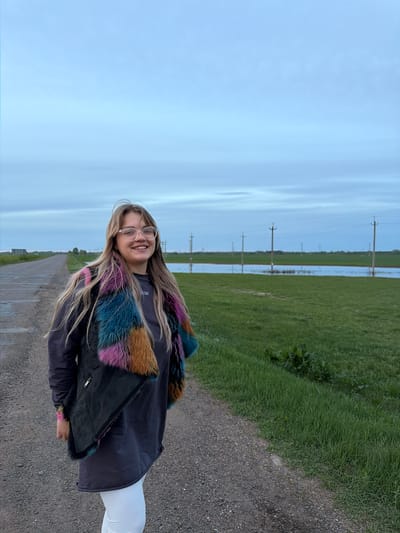 A medium shot depicts a young woman standing on a gravel path in Azyakul', Russia. She is wearing glasses, a dark jacket with colorful faux fur trim, and white leggings. The path stretches into the distance, flanked by a grassy field on one side and a paved road on the other. In the background, utility poles are visible across a flooded area, suggesting recent heavy rainfall. The sky is overcast. The woman is smiling at the camera.