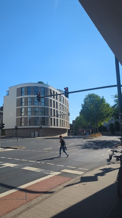 In a sunlit urban environment in Fulda, Germany, a woman with dark hair is captured mid-stride, crossing a multi-lane intersection on a pedestrian crossing. She is wearing a dark backpack and blue jeans, and her right arm is raised to her ear as if speaking on her phone. The traffic lights above the intersection are red, indicating a stop for vehicular traffic. A modern, curved building with numerous windows and vertical louvers dominates the left side of the frame, its white facade contrasting with the blue sky. To the right, a large, green tree provides a natural element, and further in the background, other buildings and streetlights are visible. Bicycles are parked near the sidewalk on the right side of the image, their shadows cast across the pavement. The scene depicts a typical moment of urban life, with elements of both infrastructure and pedestrian activity under a clear, bright sky.