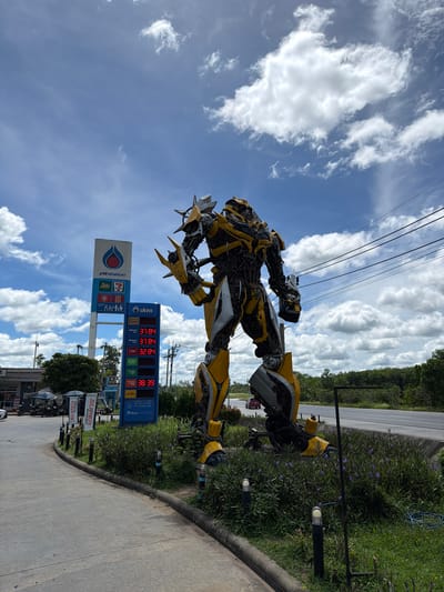 A large, yellow and gray robot statue, resembling Bumblebee from the Transformers franchise, stands in front of a PTT gas station and a 7-Eleven convenience store on a sunny day in Tha Ruea, Thailand. The robot is positioned on a grassy area with some small purple flowers. A PTT gas price sign displaying fuel prices in Thai baht is to the left of the robot. The sky is blue with scattered white clouds, and power lines are visible above. The scene appears to be along a road, with a few vehicles in the background. The robot statue is the dominant element in the foreground, with the gas station and convenience store providing context for the location. There are no people directly interacting with the robot.