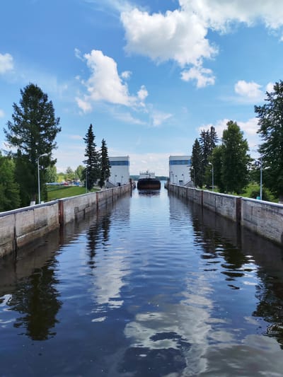 The image depicts a view from within a canal lock, looking towards the open water and a ship. The canal is flanked by concrete walls with mooring posts. On the right side, there is a white building with blue accents, and trees line the bank. On the left side, similar concrete walls are visible, with tall evergreen trees and lampposts. In the distance, a ship is positioned in the waterway, approaching what appear to be lock gates or a dam. The sky is blue with scattered white clouds, and the water reflects the sky and surrounding trees. The scene is set in the city of Белоусово, Russia, and captures a functional part of the waterway system.