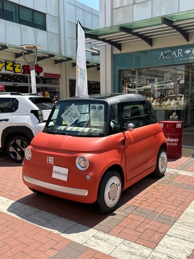 The image depicts an outdoor pedestrian commercial area in Edirne Merkez, Turkey, under clear daylight. A small, matte red Fiat Topolino electric vehicle is prominently parked on reddish-brown brick pavers, which are interspersed with lighter-colored rectangular sections. The Topolino features a black soft-top roof, circular chrome-colored hubcaps, and a white sign on its windshield displaying "200.000 TL 12 AY %0 FAİZ" (200,000 Turkish Lira, 12 months, 0% interest). A smaller white sign on its front bumper reads "TOPOLINO."

To the left and slightly behind the Topolino, the rear section of a white SUV, possibly another Fiat model, is visible. Its wheels also bear the "FIAT" logo.

The background consists of commercial buildings. On the left, a multi-story building with a light-paneled facade features a store with a partially visible yellow and red logo, reading "YZZ SHOP." A modern street lamp with a circular luminaire is positioned in front of this building. Between the left building and the right building, a tall, white vertical flag with a multi-colored "FIAT" logo stands.

On the right, a building with a large glass storefront houses a store named "ACAR" with "since 199..." visible below its name, displaying jewelry and accessories. A green-tinted glass canopy with metal supports extends from the building facade above this store. To the right of the red Topolino, a red barrel featuring "FIAT," "akıncı," and a phone number "235 52 80" is placed on the pavers. One person is partially visible behind the white SUV, looking towards the shops.