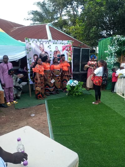 This image captures an outdoor celebratory event in the city of Amokvo, Nigeria. The scene is set against a backdrop featuring a building with a reddish-brown corrugated metal roof and lush green trees. To the left, a white canopy tent is visible.
A prominent vertical banner displaying "CELEBRATION OF LOVE" and multiple photographic portraits of individuals serves as a central feature. Positioned in front of this banner are five women, uniformly dressed in traditional Nigerian attire consisting of bright orange off-shoulder tops with ruffled sleeves and ankle-length skirts with geometric patterns in black, orange, and white. Each woman carries a small purse or clutch, and they are posing for a photograph.
Immediately to the right of the banner, two large black speaker units are stacked. A section of artificial green turf is laid on the ground, creating a designated area in front of the banner. A cluster of artificial blue and white flowers with green foliage is placed on this turf.
A young woman with long dark hair, wearing a white t-shirt, black leggings, and a red patterned wrapper tied around her waist, stands barefoot on the artificial turf, facing the group of women and the banner. She is holding a small dark object, likely a mobile phone or camera, directed towards them, indicating she is capturing an image or video. Behind her, a woman in a pink and beige traditional outfit with a blue headscarf is partially visible, holding what appears to be a traditional fan or whisk. A young boy in a red shirt is partially visible on the far right, near a white decorative structure adorned with artificial flowers and green vines.
To the far left, a man in a light purple long-sleeved traditional outfit and yellow open-toed slippers stands. Other individuals are seated under the tent structure in the background. In the immediate foreground, a white table with a light-colored cloth is partially visible, where a person's dark-skinned hand is holding two small clear plastic bottles with purple caps. The ground surrounding the artificial turf is exposed dirt.