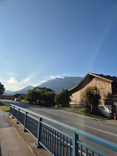 The image depicts a rural scene in Niederndorf, Austria, on a clear day with a bright blue sky. In the foreground, a dark blue metal railing with horizontal bars runs along the edge of a paved walkway. Adjacent to the walkway is a road with a white dashed line marking the lanes, and a narrow strip of grass and trees separates the road from a wooden fence.
Further into the scene, several buildings are visible. On the right, a large, rustic wooden building with a gabled roof covered in wooden shingles dominates the view. A small sign is affixed to its side. To the left of this building, a smaller wooden structure with a pitched roof can be seen. Behind these structures, trees with green foliage are scattered, and further in the background, rolling hills and imposing mountains rise against the sky.
A white car is parked on the side of the road in front of some trees. The overall impression is one of a tranquil, sunlit rural landscape.