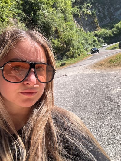A close-up, selfie-style portrait of a young woman with blonde hair and large, orange-tinted sunglasses is shown. She is positioned on the left side of the frame, her face taking up a significant portion of the foreground. Behind her, a winding asphalt road recedes into the distance, bordered by steep, wooded hillsides. Several cars are visible on the road, appearing to be in motion. The environment suggests a sunny day in a rural or mountainous area, possibly a scenic route in L'Essard, France, given the presence of the road and natural landscape. The woman's expression is neutral.