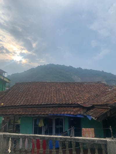 The image shows a residential dwelling with a terracotta tile roof located in Lagadar, Indonesia (latitude: -6.928, longitude: 107.535). The house is a single-story structure with a teal green exterior, a gated balcony, and a section of an adjacent structure visible to the right. Clothes are hung out to dry on a line extending from the house towards the left edge of the frame. The home faces a verdant hillside which is obscured by haze in the distance. The sky is overcast and light gray with thin clouds. The sun is hidden behind the clouds, resulting in a low-light, muted color scheme.