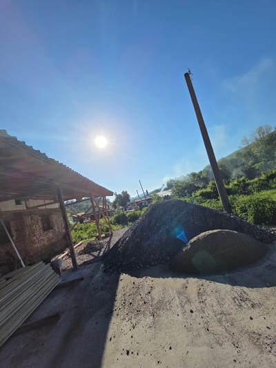 The image depicts a scene in Kaleytsa, Bulgaria, oriented with the sky dominating the upper portion. A wooden utility pole stands against the blue sky. In the background, there is green foliage and what appears to be smoke rising. In the foreground there is a concrete embankment, gravel, and an old brick building undergoing construction. Lumber lies near the building. The sun shines brightly.