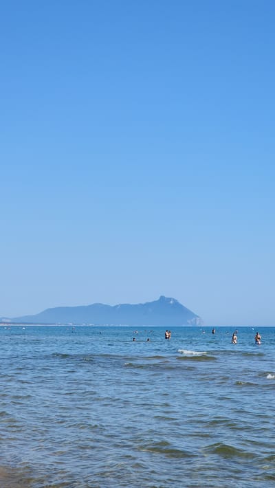 The image depicts a bright, sunny day at a beach in Sabaudia, Italy. The sky is a clear, vibrant blue, taking up the majority of the frame. Below the sky, a calm, turquoise sea stretches across the horizon. In the distance, a prominent mountain range is visible, its slopes appearing green and blue under the bright sunlight. The water is populated by numerous individuals, some swimming, others standing and socializing. The waves are gentle, creating small ripples on the surface. The overall scene evokes a sense of a leisurely summer day at the seaside.